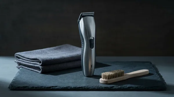 A plain unbranded brushed-steel cordless beard trimmer standing upright on a dark slate slab, beside a folded charcoal towel and a small wooden cleaning brush